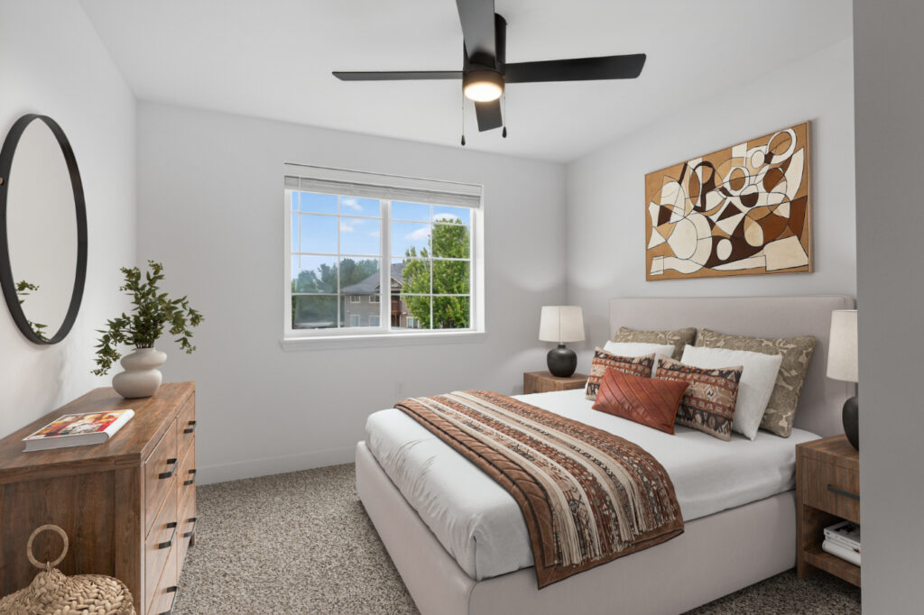 Modern bedroom with a bed, patterned blanket, pillows, wooden dresser, nightstands, lamps, round mirror, abstract wall art, and a ceiling fan. Large window lets in natural light, showing trees and houses outside.