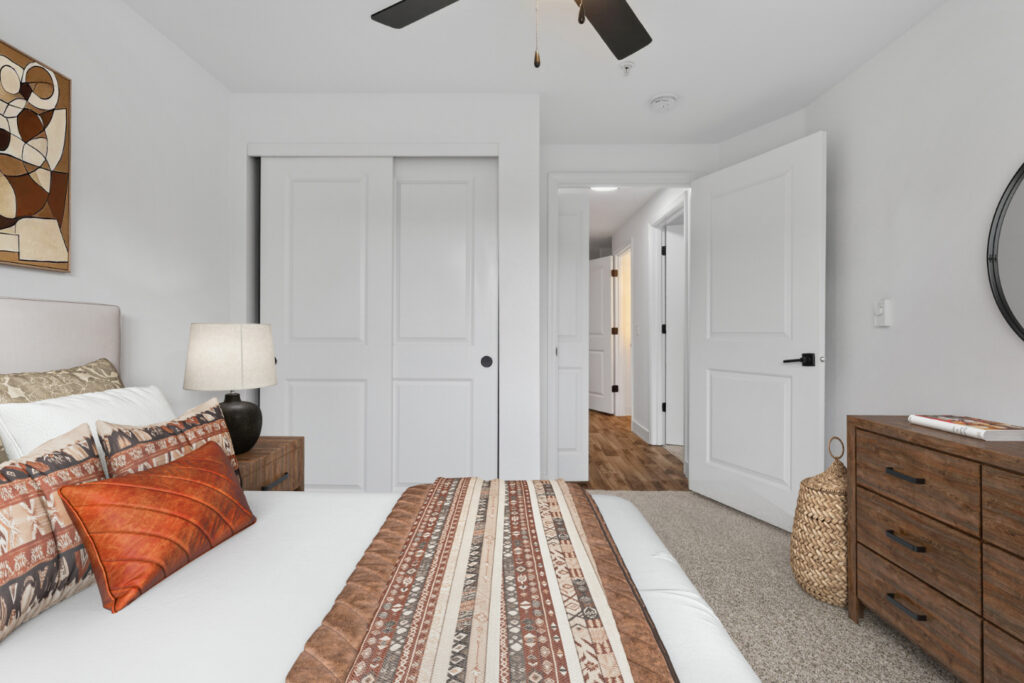 A modern bedroom with a bed in the foreground, patterned bedding, and orange pillows. A wooden dresser and a lamp are on the left, with double closet doors and an open door to a hallway in the background.