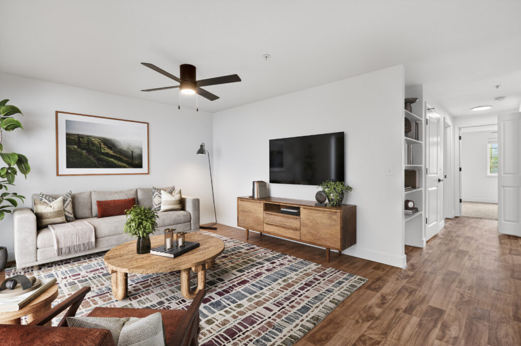 Modern living room with a gray sofa, wood coffee table, TV on a wooden stand, patterned rug, plants, and wall art. White walls and wood flooring give a clean, airy feel. Hallway is visible on the right.