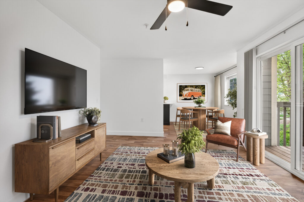 Modern living room with wooden furniture, TV mounted on the wall, round coffee table, patterned rug, and large windows letting in natural light; dining area visible in the background.
