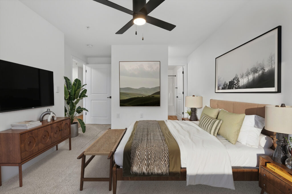Modern bedroom with a large bed, green pillows, wood furniture, a bench, and two landscape photos on white walls. Ceiling fan, TV, lamp, and a potted plant add to the cozy, minimalist decor.